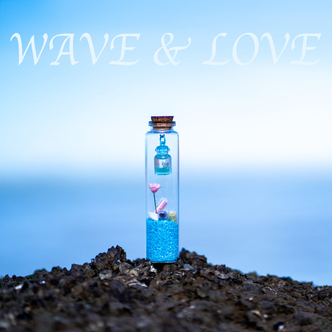 The WAVE & LOVE personalized keepsake bottle, a unique message in a bottle gift, rests on a dark, rocky shore against a serene, blurred blue ocean background. Inside the glass bottle, vibrant blue sand holds a tiny pink flower, a seashell, and colorful message capsules, with a miniature bottle hanging from the cork.