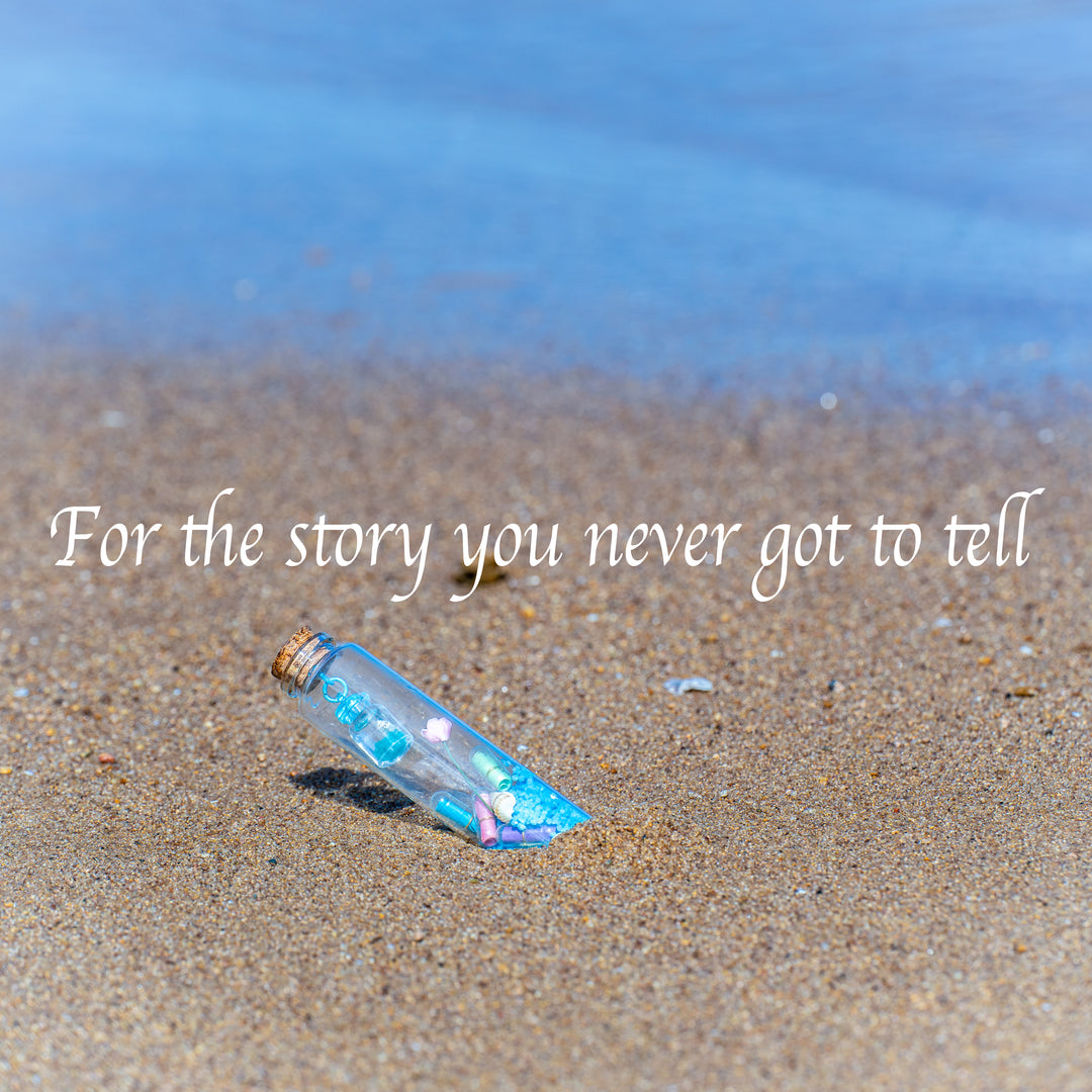 A personalized message in a bottle gift lies half-buried in wet sand at the water's edge, with the blue ocean in the background. The image text reads, "For the story you never got to tell"