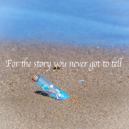 A personalized message in a bottle gift lies half-buried in wet sand at the water's edge, with the blue ocean in the background. The image text reads, "For the story you never got to tell"